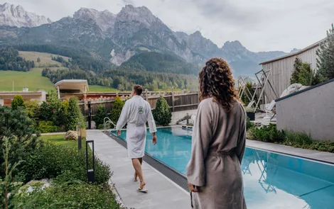 Herbstzauber & Wellnesstage Paar im Bademantel geht am Außenpool eines Wellnesshotels in den Alpen entlang